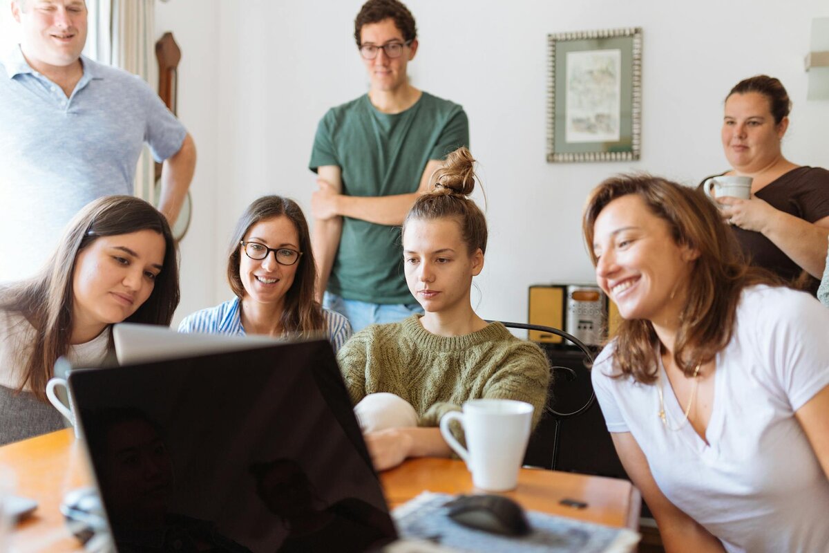 A group of seven people gathers around a table, looking at a laptop screen together in a casual indoor setting.