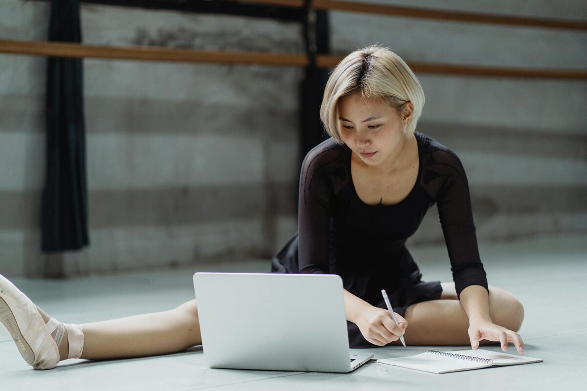 A dancer in a leotard and ballet shoes sitting on a studio floor, writing in a notebook beside an open laptop.