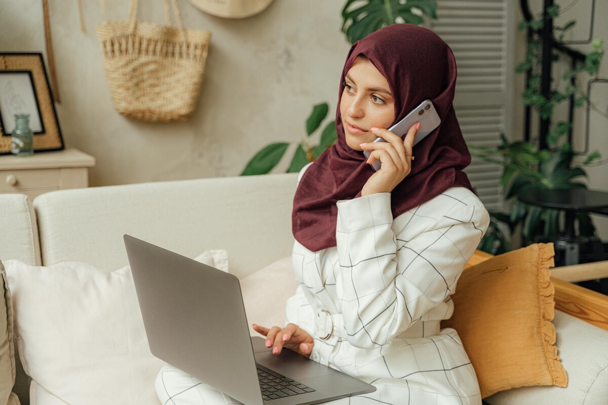 woman wearing maroon headscarf talking on the phone in front of her laptop