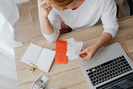 Overhead view of a woman reviewing receipts at a desk with a laptop, notebook, and cash.