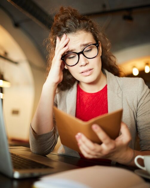 Stressed woman with glasses reading notebook near laptop