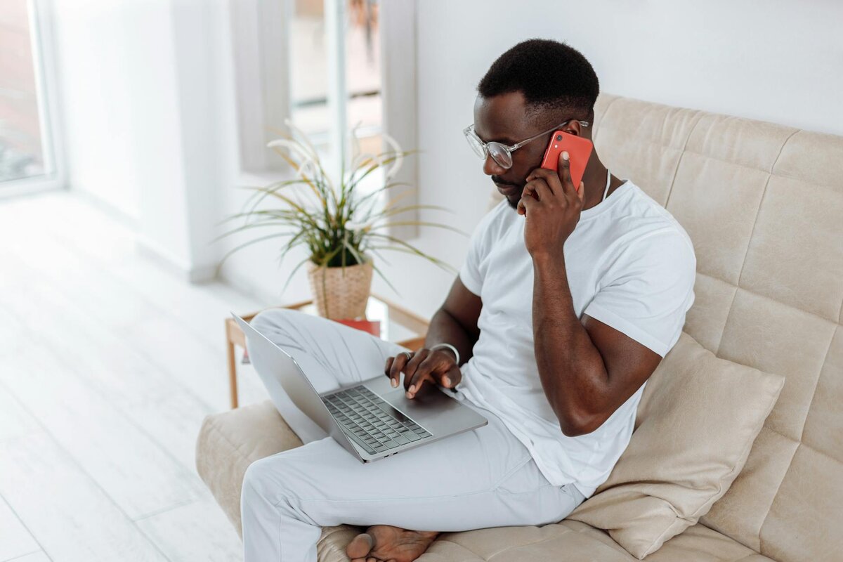 A man sitting on a couch, working on a laptop while talking on a phone call