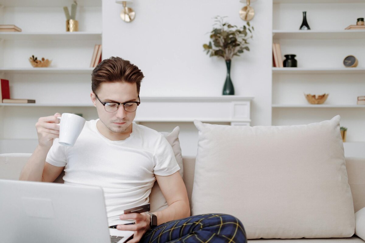 Man on a sofa holding a card and mug while using a laptop
