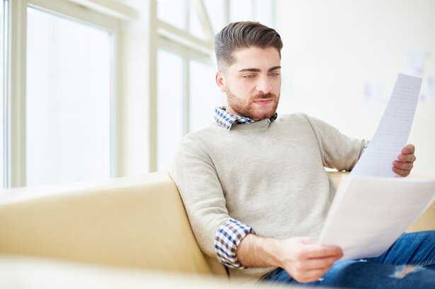 Young man sitting on sofa carefully reading printed documents