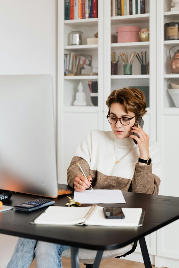 Woman wearing glasses sits at a desk talking on the phone while writing notes, with a desktop computer and bookshelf in the background.