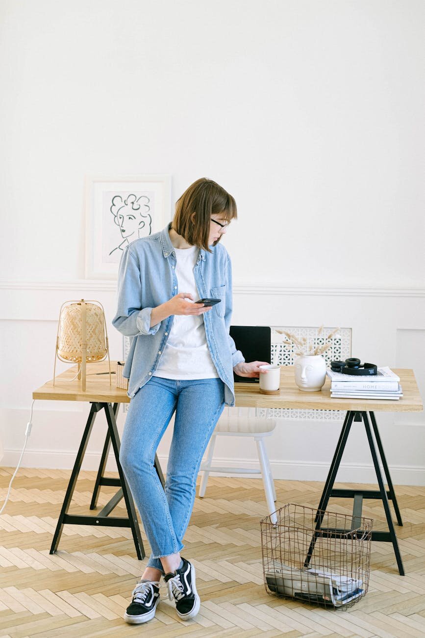 a woman leaning on a desk, holding a phone and mug in a bright home office with laptop and books.