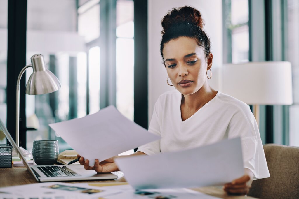 Woman looking concentrated while reviewing paperwork at a desk with a laptop.