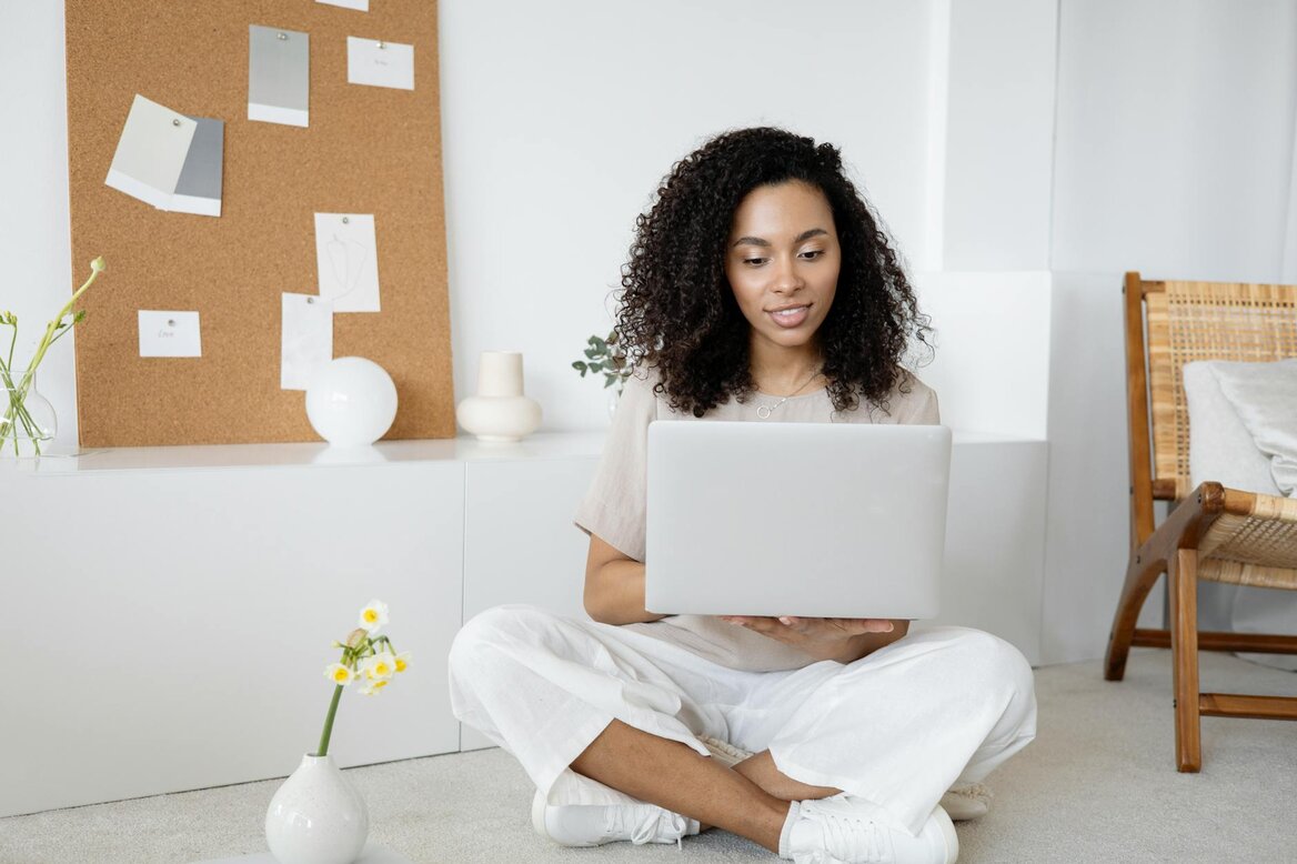 Woman sitting cross-legged on the floor using a laptop, with a corkboard and flowers nearby