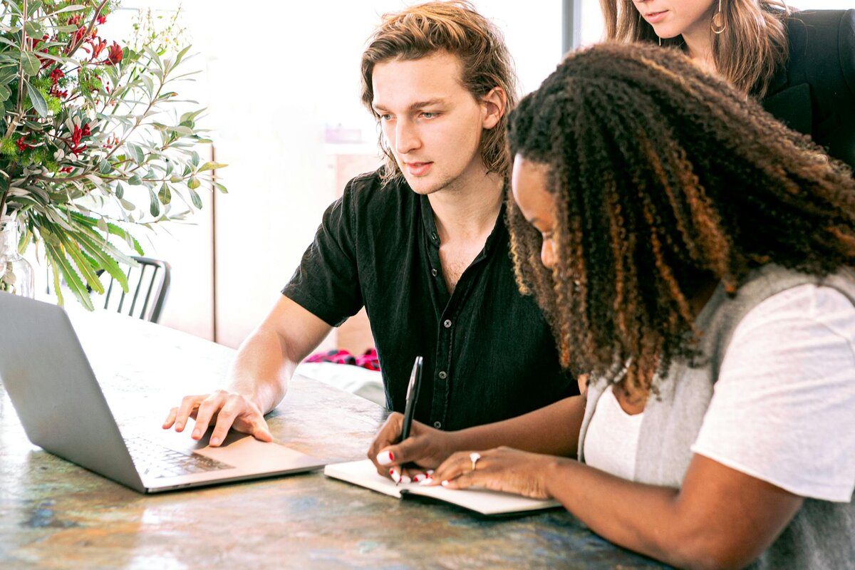 Three people collaborate at a table, with a man pointing at a laptop while a woman takes notes in a notebook nearby.