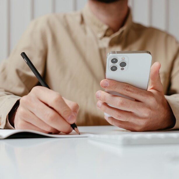 Close-up of a man holding a smartphone in one hand while writing with a pencil in a notebook with the other.