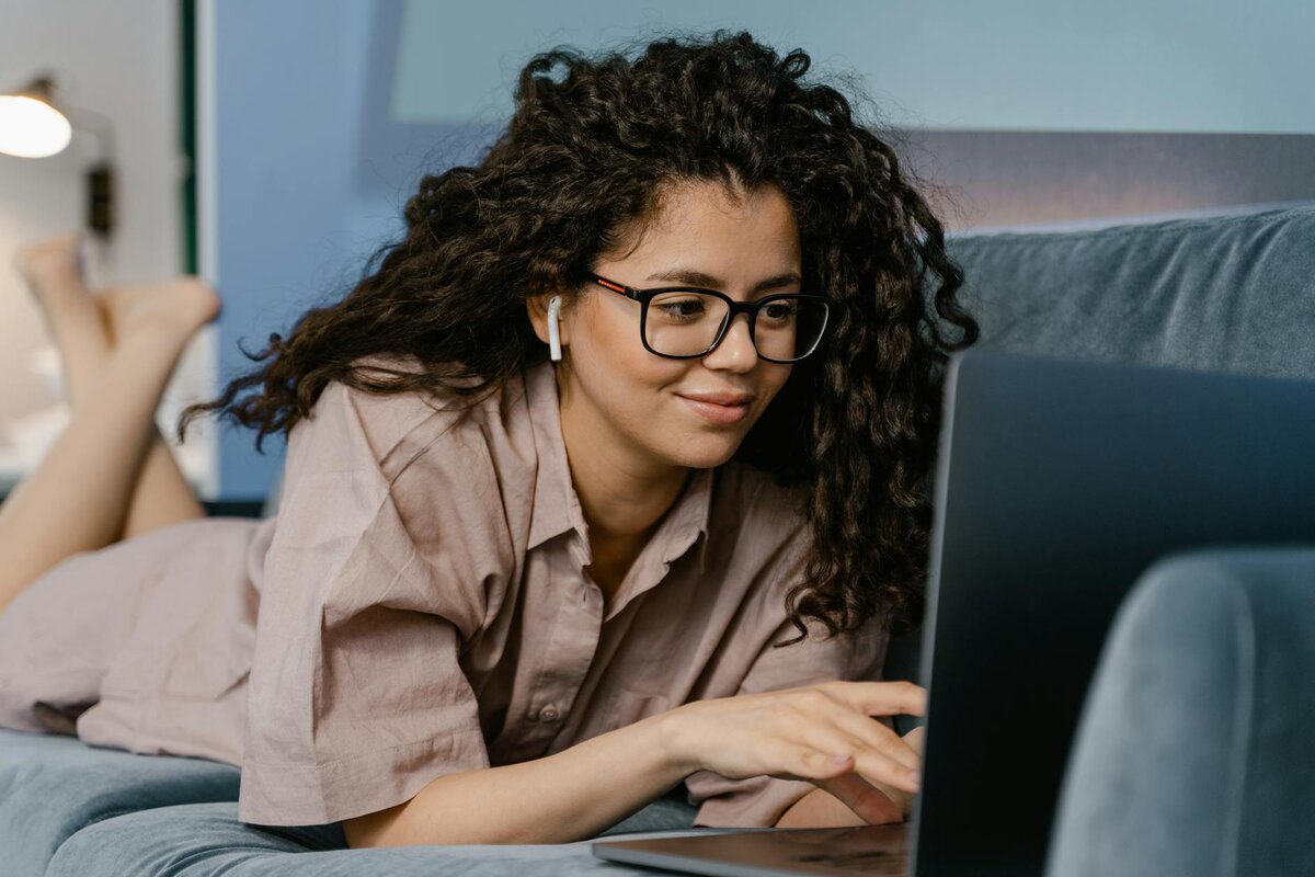 woman with curly hair and glasses lies on a couch, smiling as she types on a laptop with an earbud in one ear.