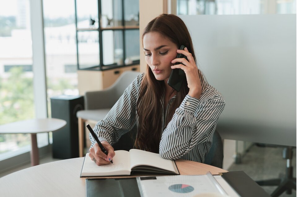 Self-employed woman on a phone call while taking notes and reviewing finances