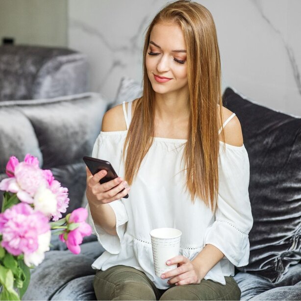 Smiling woman sitting on a velvet couch, looking at her phone while holding a coffee cup, with pink flowers nearby.