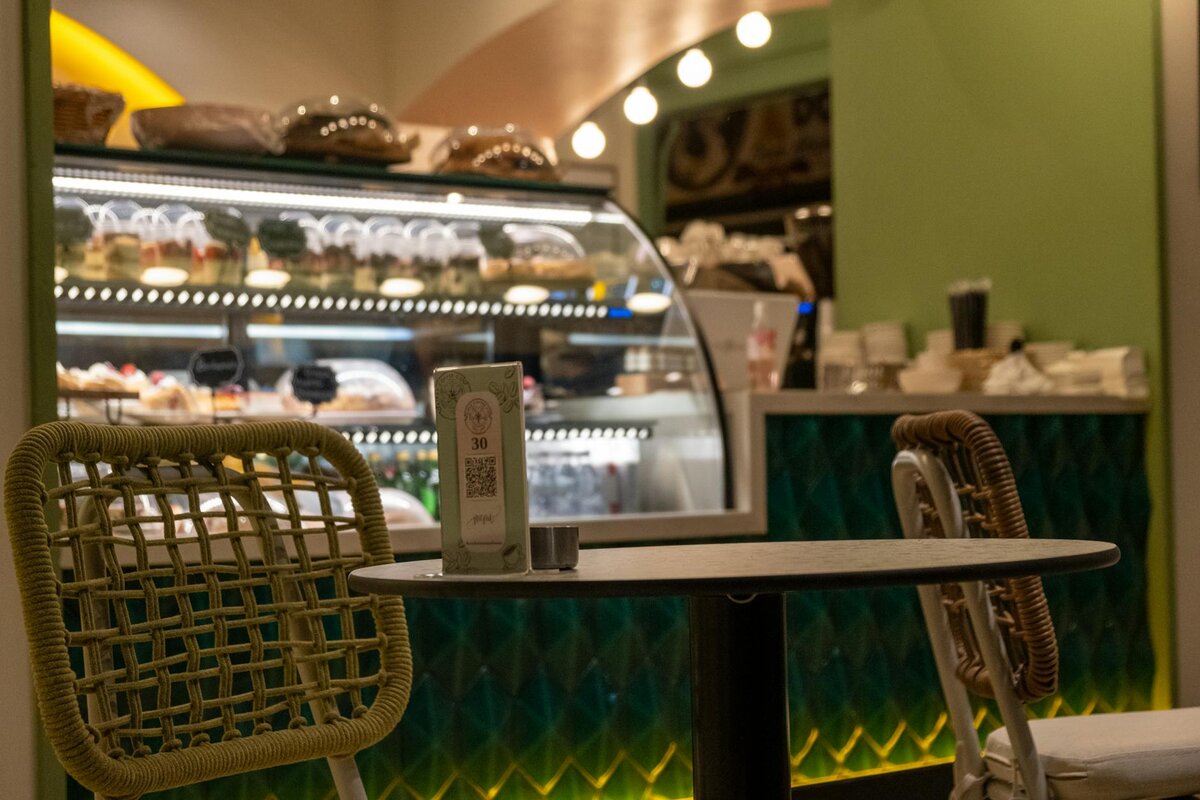 An empty café table with a table number card and QR code, set in front of a glass pastry display case in a green-themed bakery.