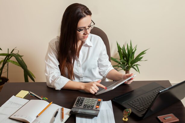 Woman wearing glasses sits at a desk using a tablet, surrounded by a calculator, notebook, and laptop.