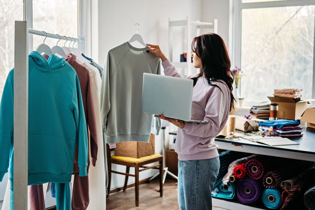 Woman holding laptop while reviewing clothing items in studio.
