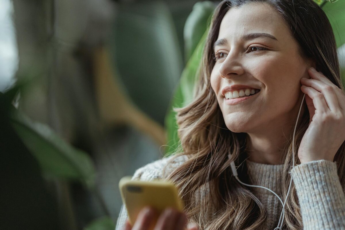 Smiling woman with earphones holding yellow smartphone near green plants
