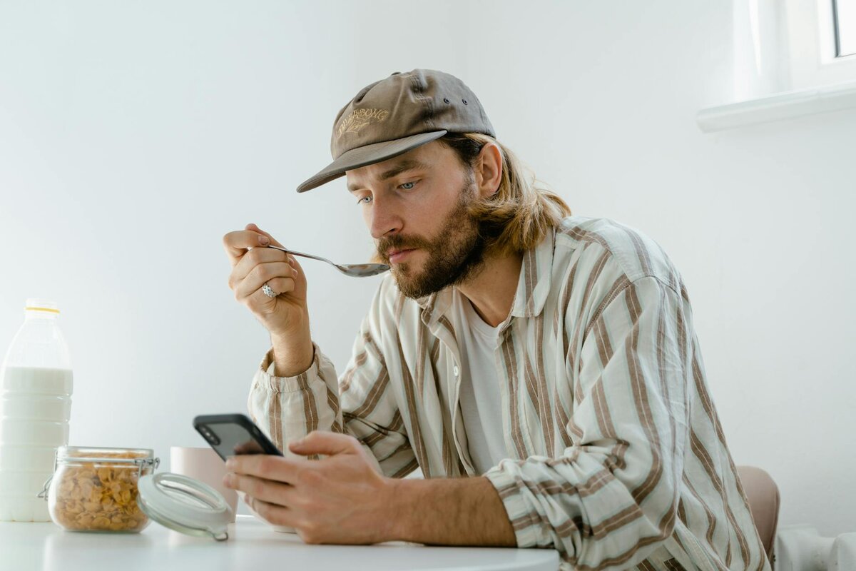 Casual man eating breakfast while browsing smartphone at home table