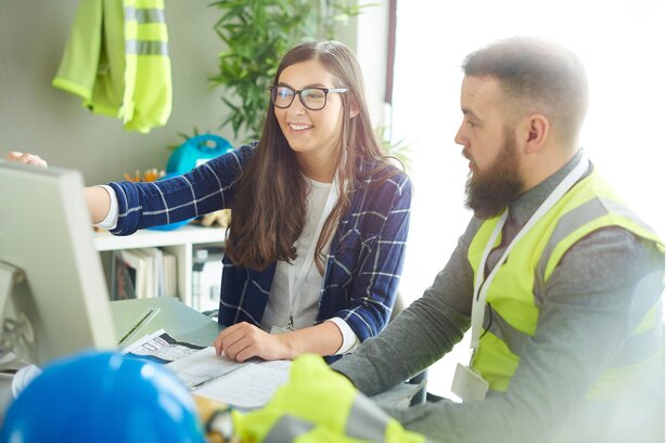 Woman in glasses pointing at a computer monitor while explaining something to a bearded man in a high-vis vest, with hard hats nearby.