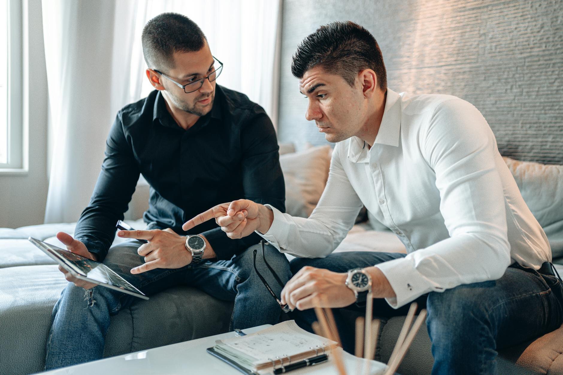 Two men sitting on a couch in discussion, one holding a tablet while the other points toward documents on a table.