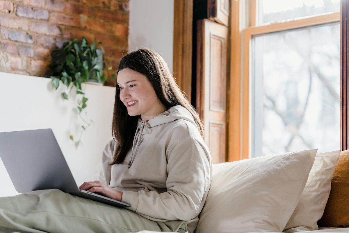 Woman sits on a bed smiling while typing on a laptop near a sunlit window.