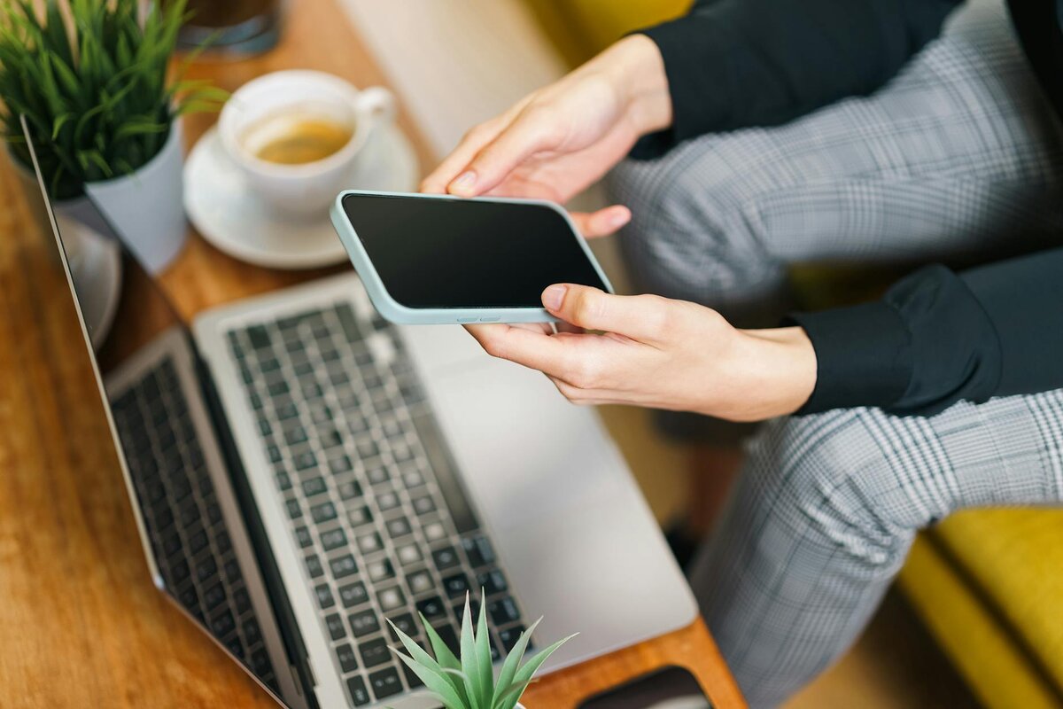 close-up overhead view of a person in plaid trousers holding a smartphone beside a laptop, with a cup of tea and small plants on the wooden desk