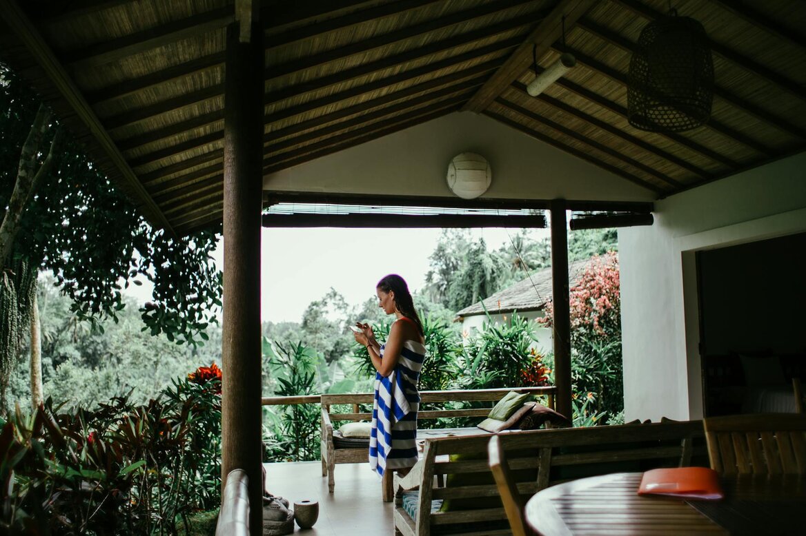 Woman stands on a covered porch looking at her phone, surrounded by lush greenery.