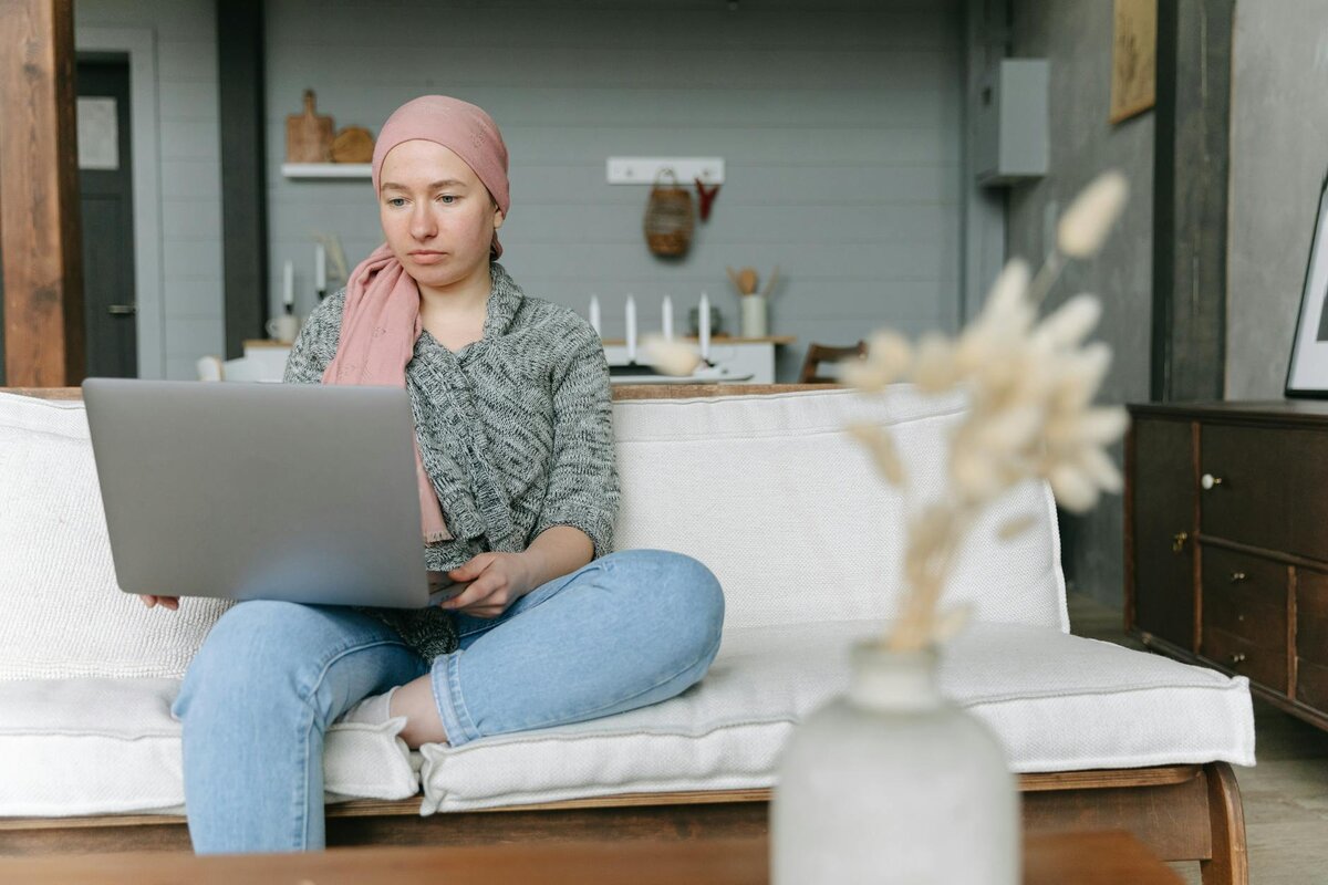 woman wearing a pink headscarf sits cross-legged on a couch, using a laptop in a cozy living room.