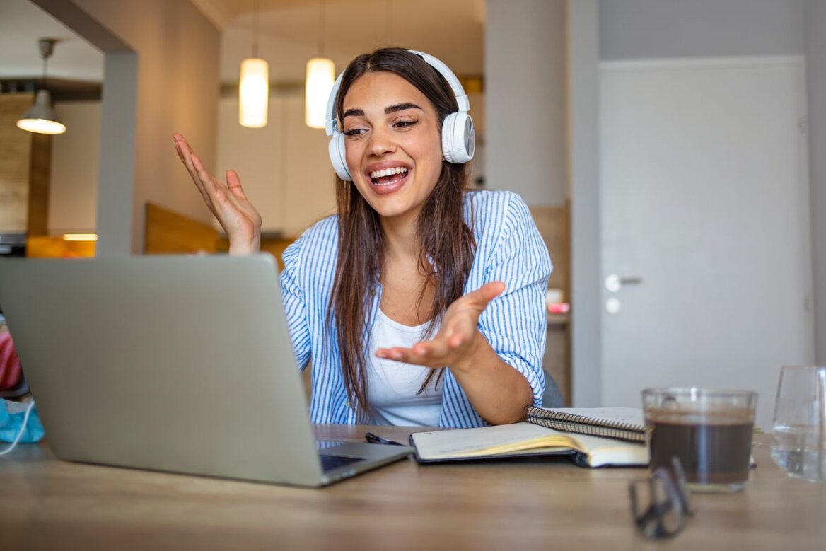 Woman wearing headphones smiles and gestures while talking in front of a laptop at a table.