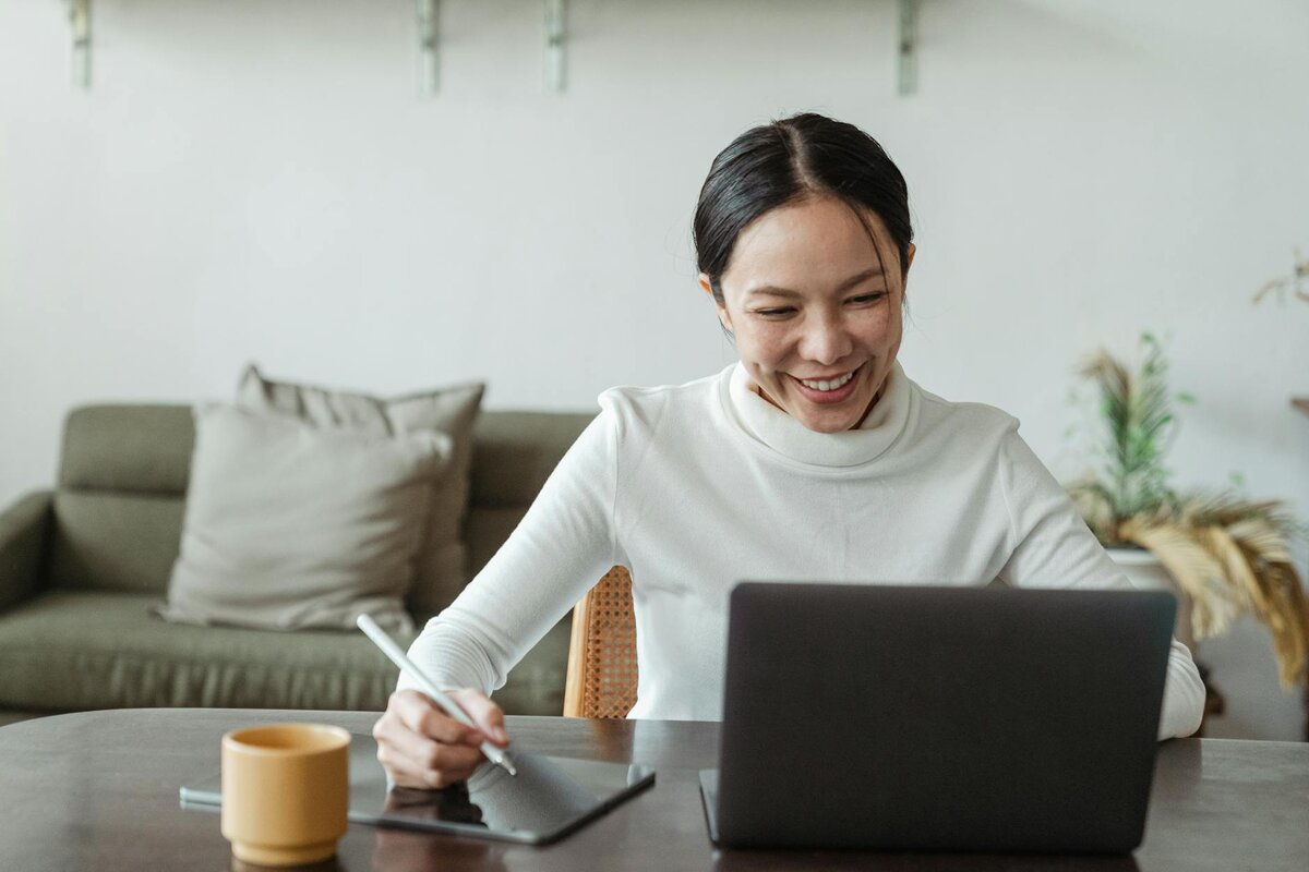 Smiling woman writing on a tablet with a stylus next to a laptop and a mug at a wooden table