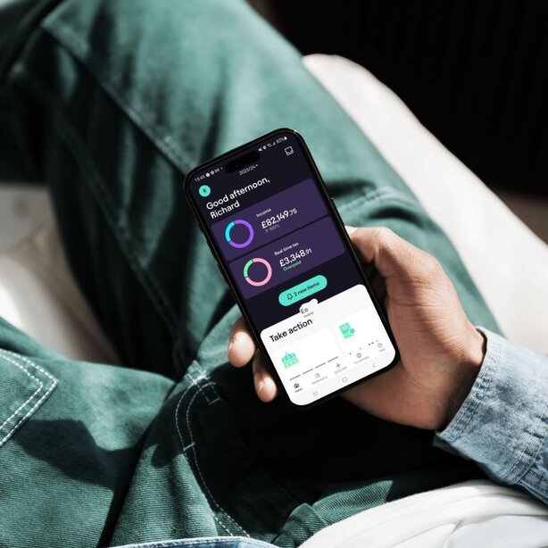 Close-up of a hand holding a phone displaying the Pie Tax app's home screen with income and tax overview, resting on a green denim-clad lap.