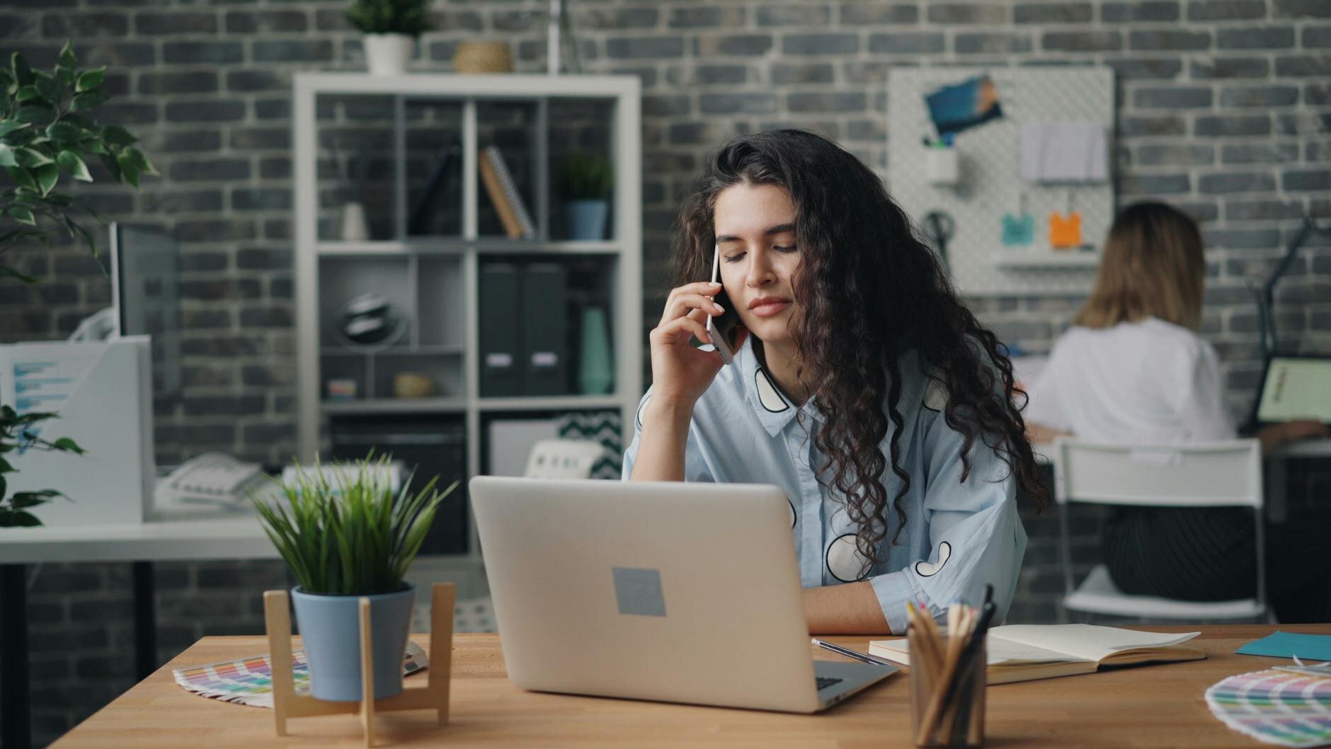 woman with long curly hair talks on her phone while sitting at a laptop in an open office, with a colleague working in the background.