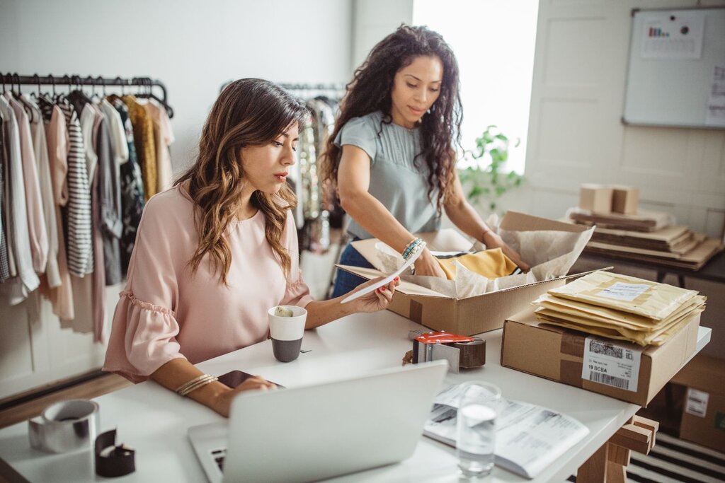 Two women working in a clothing shop, one at a laptop and the other packing items into a shipping box.