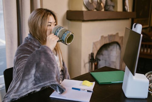 woman wrapped in a cozy blanket sips from a mug while sitting at a desk with a notebook, in front of a fireplace.