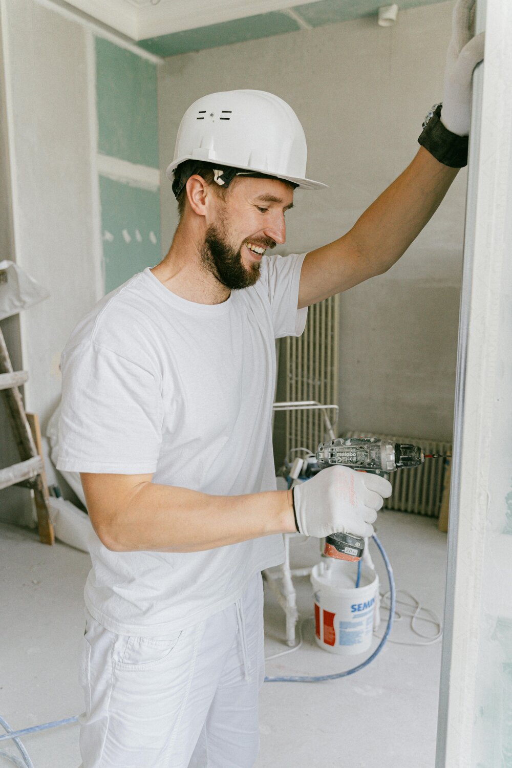 Smiling bearded man in a hard hat using a power drill at a construction site