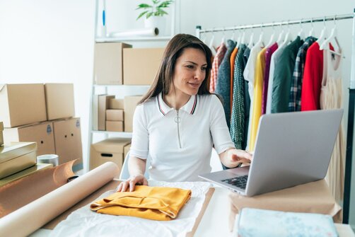 Smiling woman using a laptop at a packing table, with a clothing rack and shipping boxes behind her.