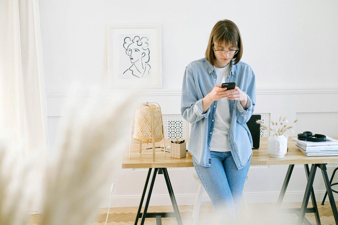 Woman wearing glasses stands using her phone near a wooden desk in a bright room.