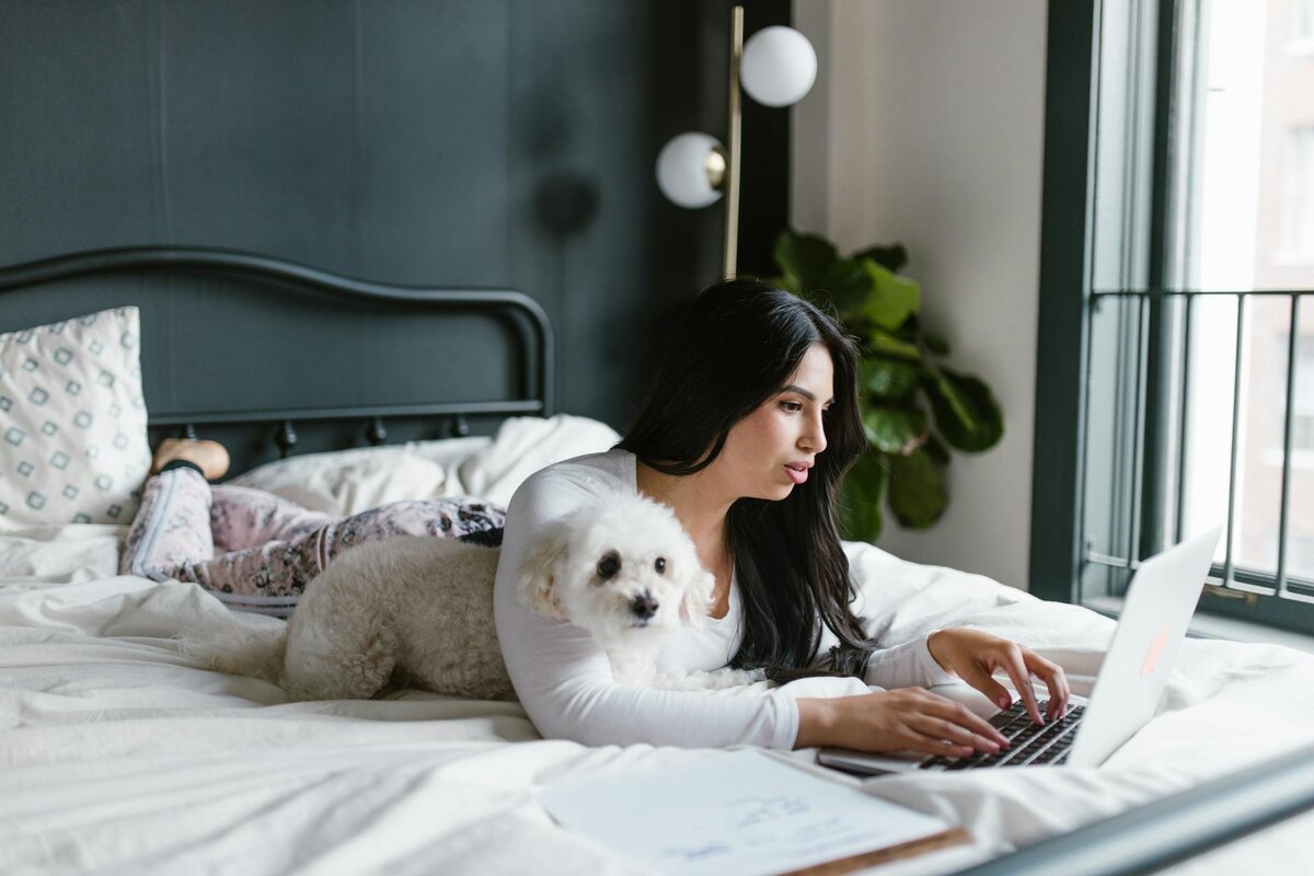 woman lying down on her bed with her dog while browsing on her laptop