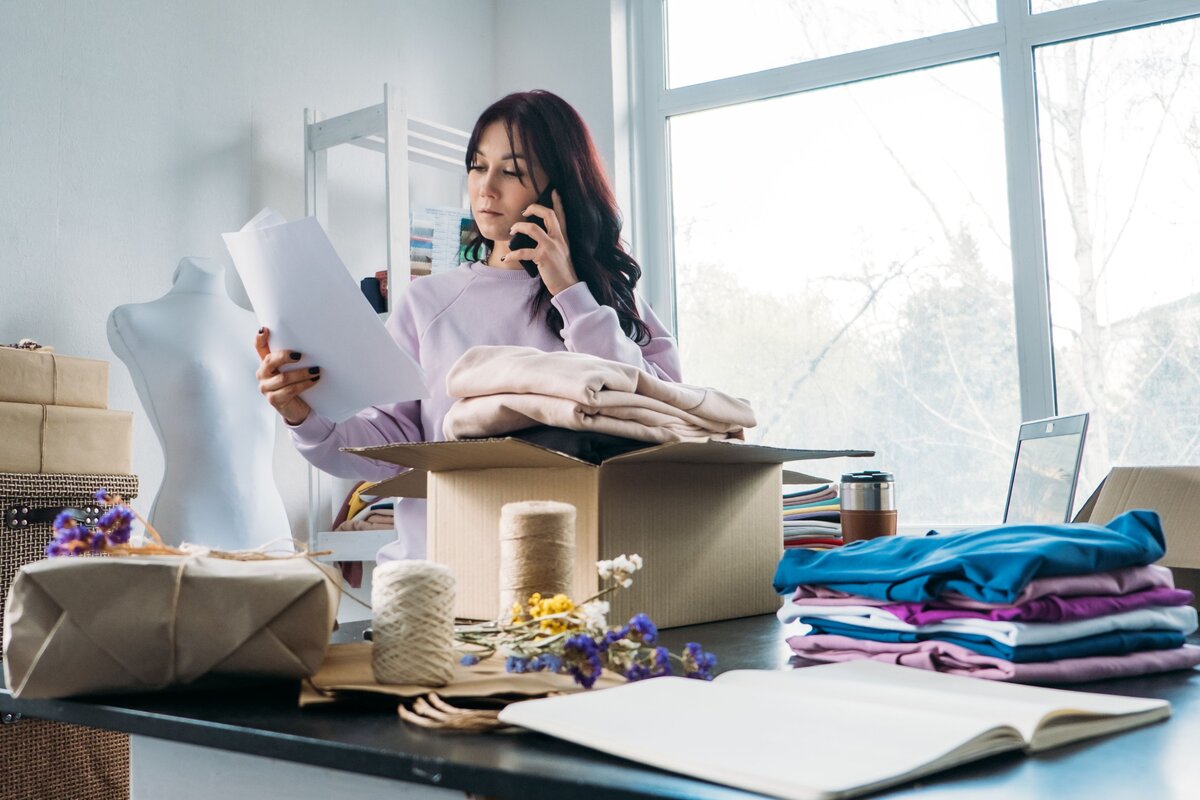 Woman talking on the phone while checking a paper and packing folded clothes into a box at a workshop table.