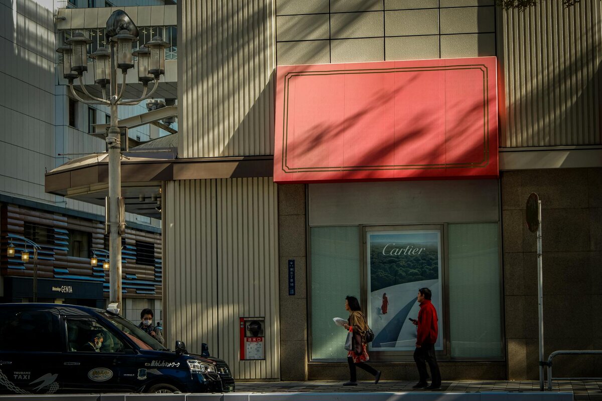 Two pedestrians walk past a Cartier storefront with a large red awning, beside a parked Tsubame taxi on a sunlit city street.