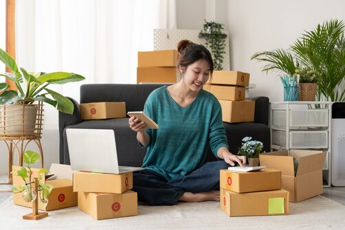 Smiling woman sits on the floor surrounded by shipping boxes, using a laptop and phone.