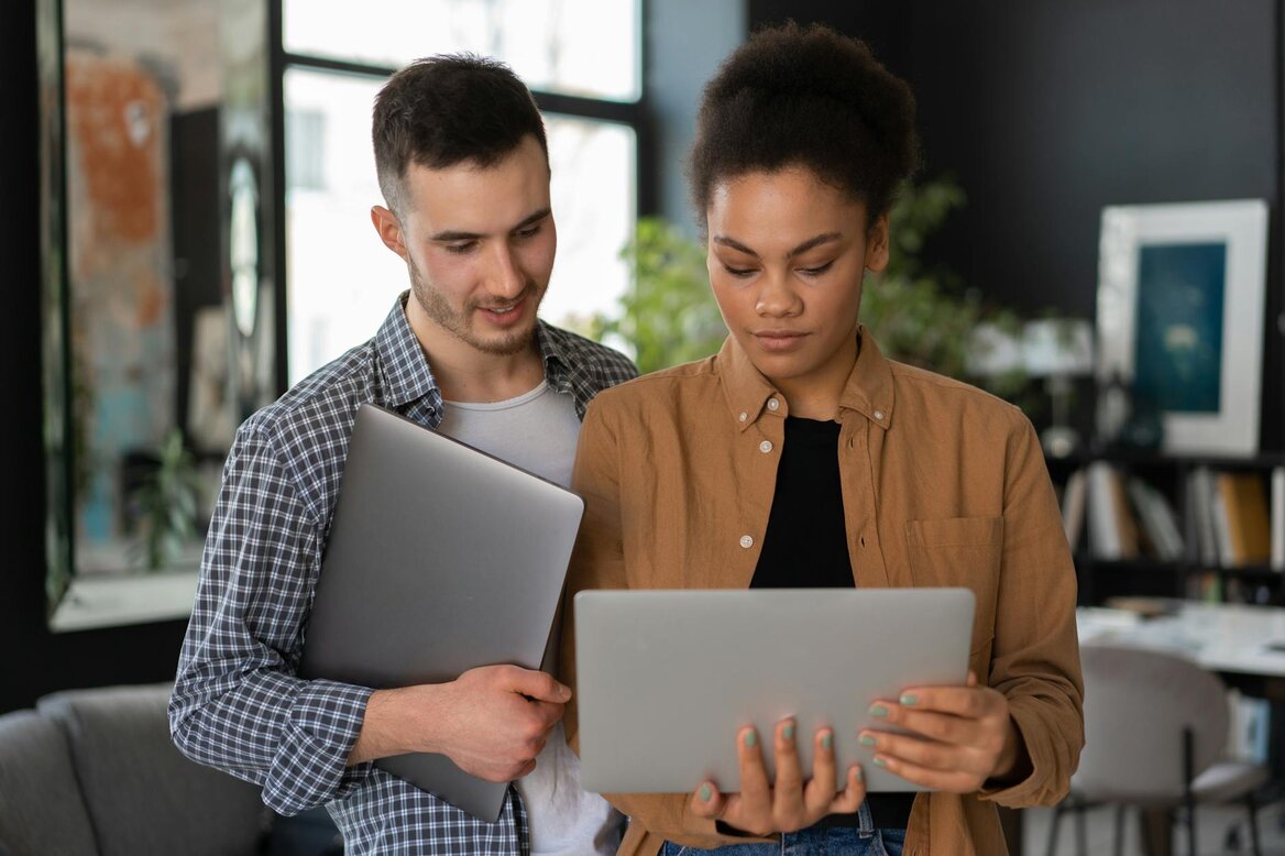 Man and woman standing together looking at a laptop screen, with the man holding a second closed laptop under his arm.Man and woman standing together looking at a laptop screen, with the man holding a second closed laptop under his arm.