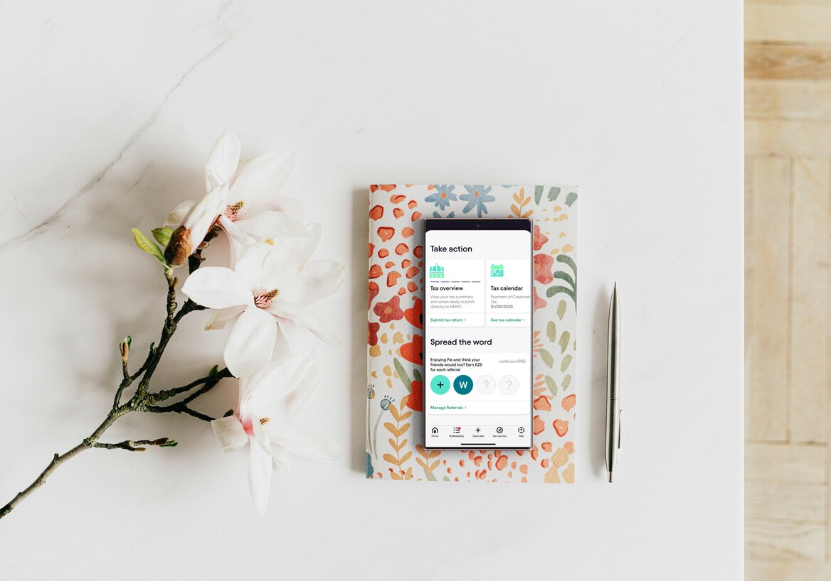 Phone showing a "Take action" app screen resting on a floral notebook, styled with a magnolia branch and a pen on a marble desk.