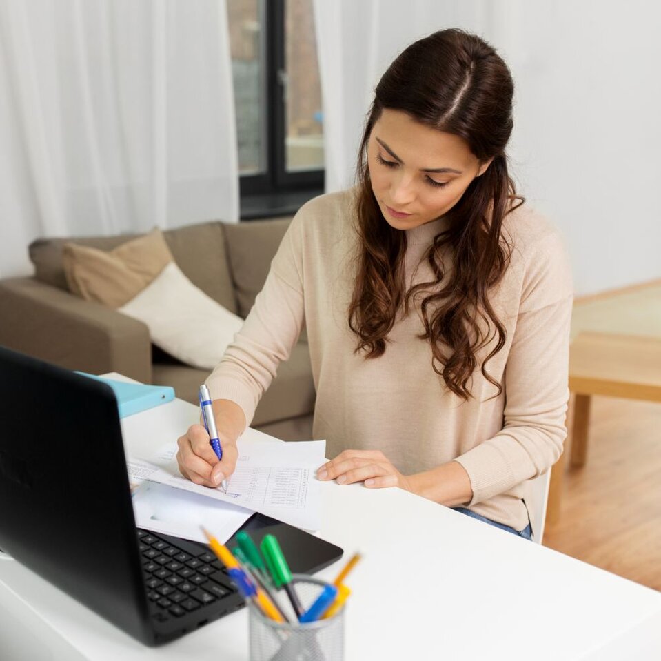 Woman writing notes at desk beside open laptop