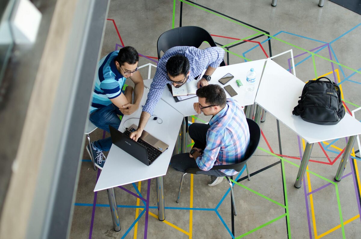 An overhead view of three men gathered around a laptop at angular white desks, set on a floor with a colorful geometric pattern