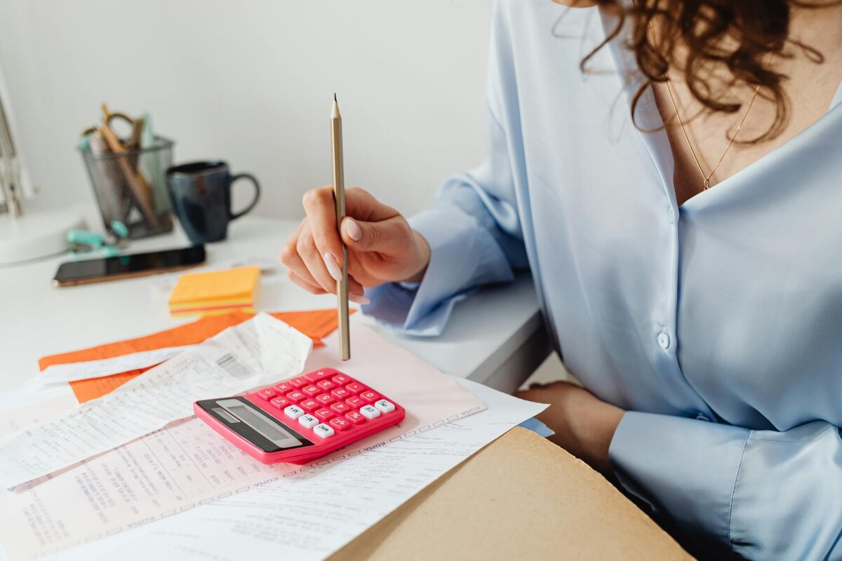 A close-up of a woman holding a pencil over a pink calculator while reviewing receipts and paperwork at her desk.