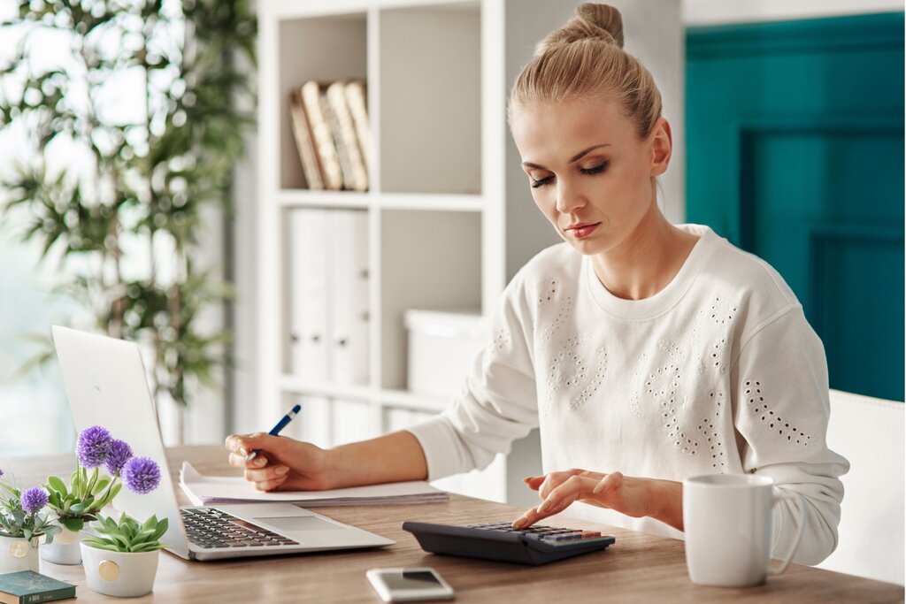Woman sits at a desk using a calculator and writing notes, with a laptop and flowers nearby.