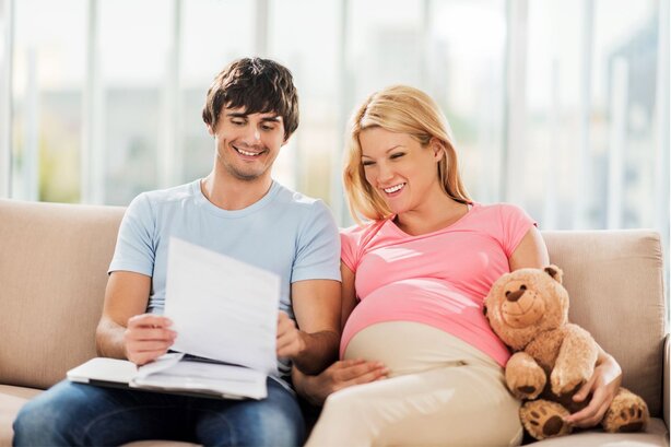 Smiling man and pregnant woman sit on a couch reviewing paperwork together, with a teddy bear beside her.
