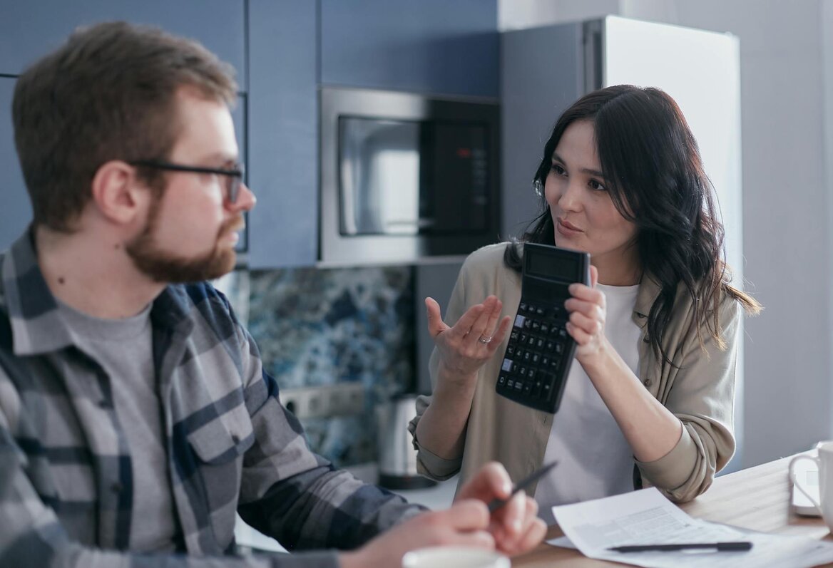 Woman showing a calculator to a man while discussing finances at a kitchen table with documents