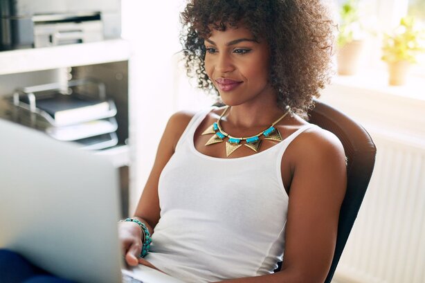 Smiling woman wearing a statement necklace, sitting at a desk looking at a laptop.